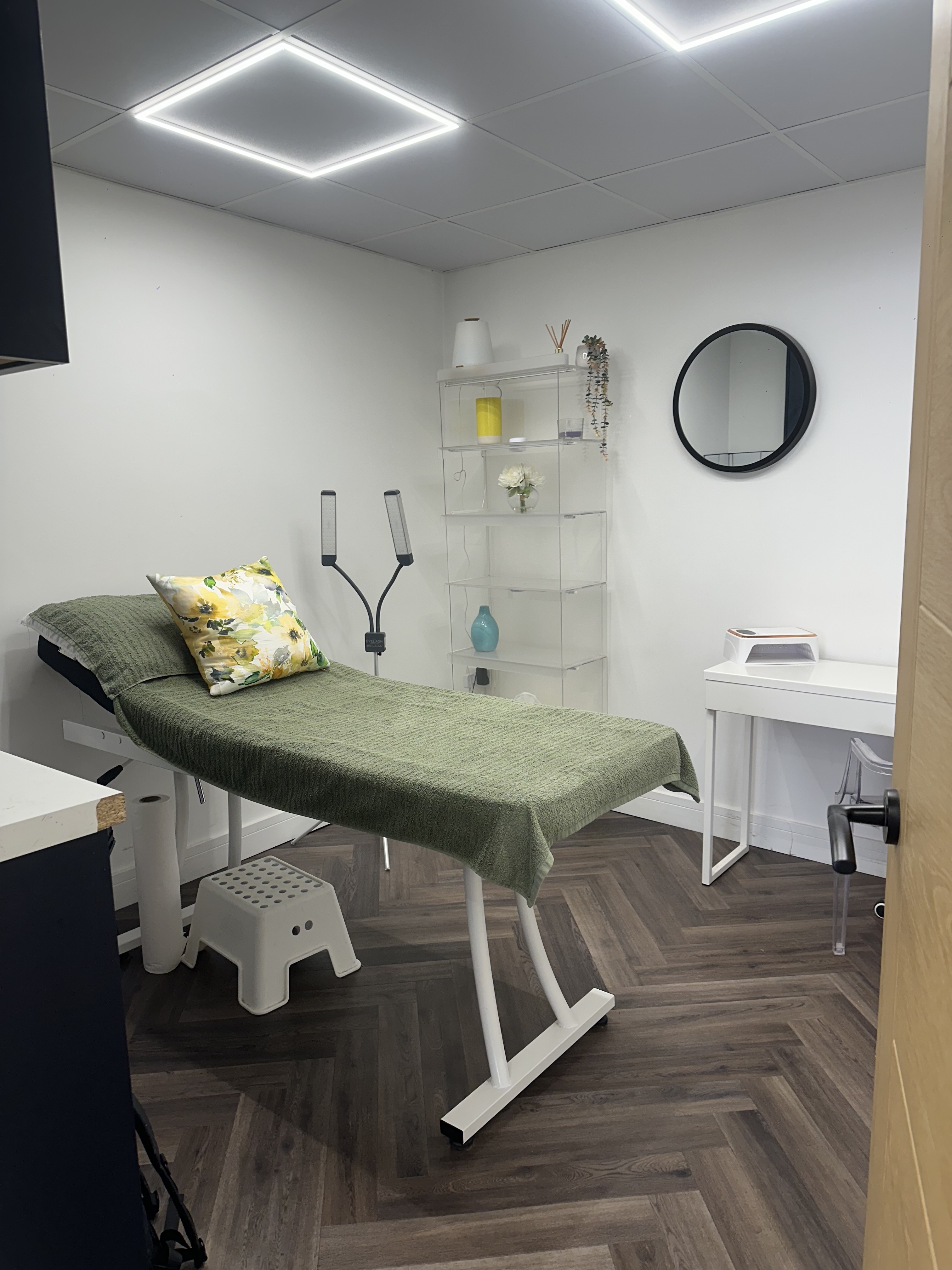The Radiant by Fran treatment room in Weston-super-Mare: a bed draped in a sage throw with a floral cushion, shelving, round mirror and nail station beneath warm LED ceiling lighting